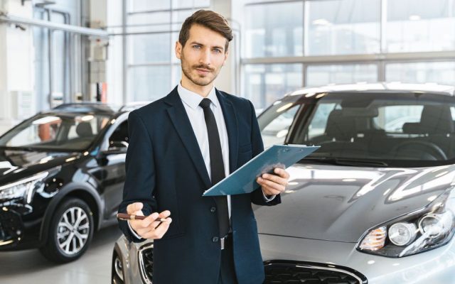Professional car dealer in business suit holding clipboard in a bright car showroom.