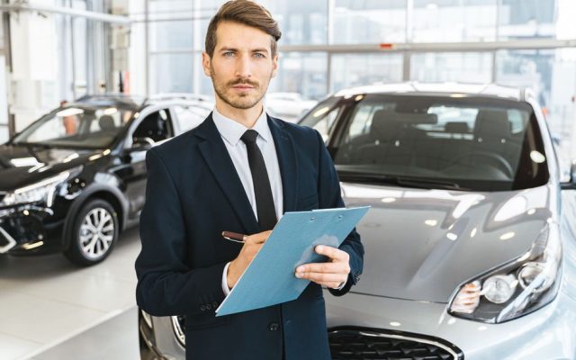 A confident car salesman in a showroom, holding a clipboard and pen.